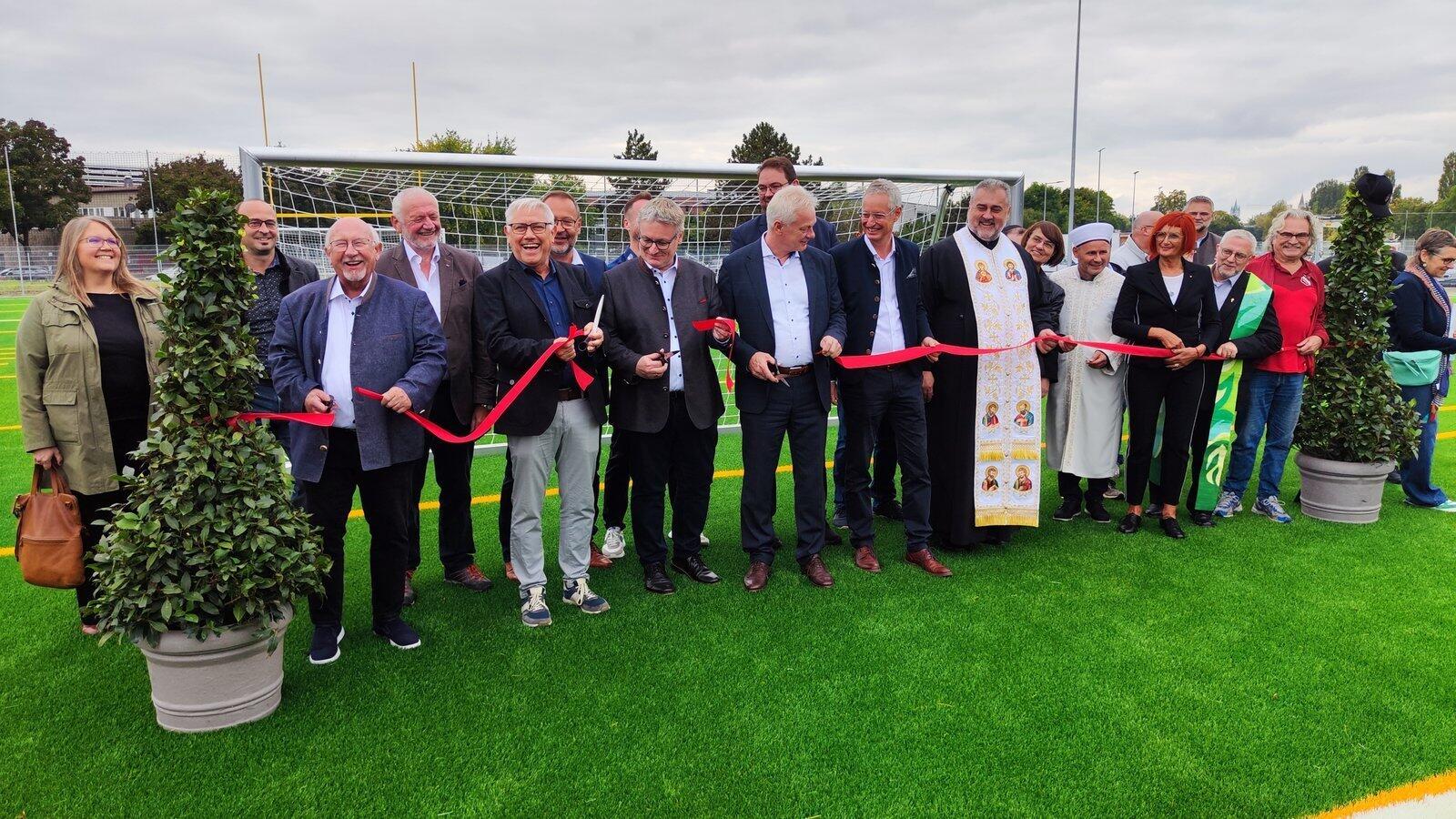 Oaspeții din sport, cler și politică, alături de ministrul federal Alois Rainer (MdB) (al 9-lea din stânga), au avut voie să taie simbolic panglica roșie și să inaugureze oficial noul teren cu gazon sintetic